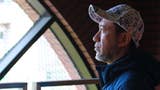 Shinji Mikami looks out of a window, turned towards the light wearing a baseball cap with upturned peak.