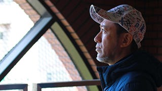 Shinji Mikami looks out of a window, turned towards the light wearing a baseball cap with upturned peak.