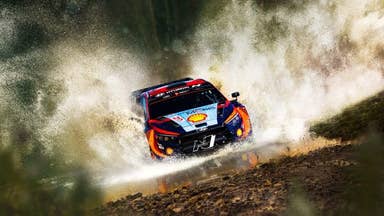A rally car with a Shell logo on its bonnet stirs up clouds of sunlit dust as it races along.