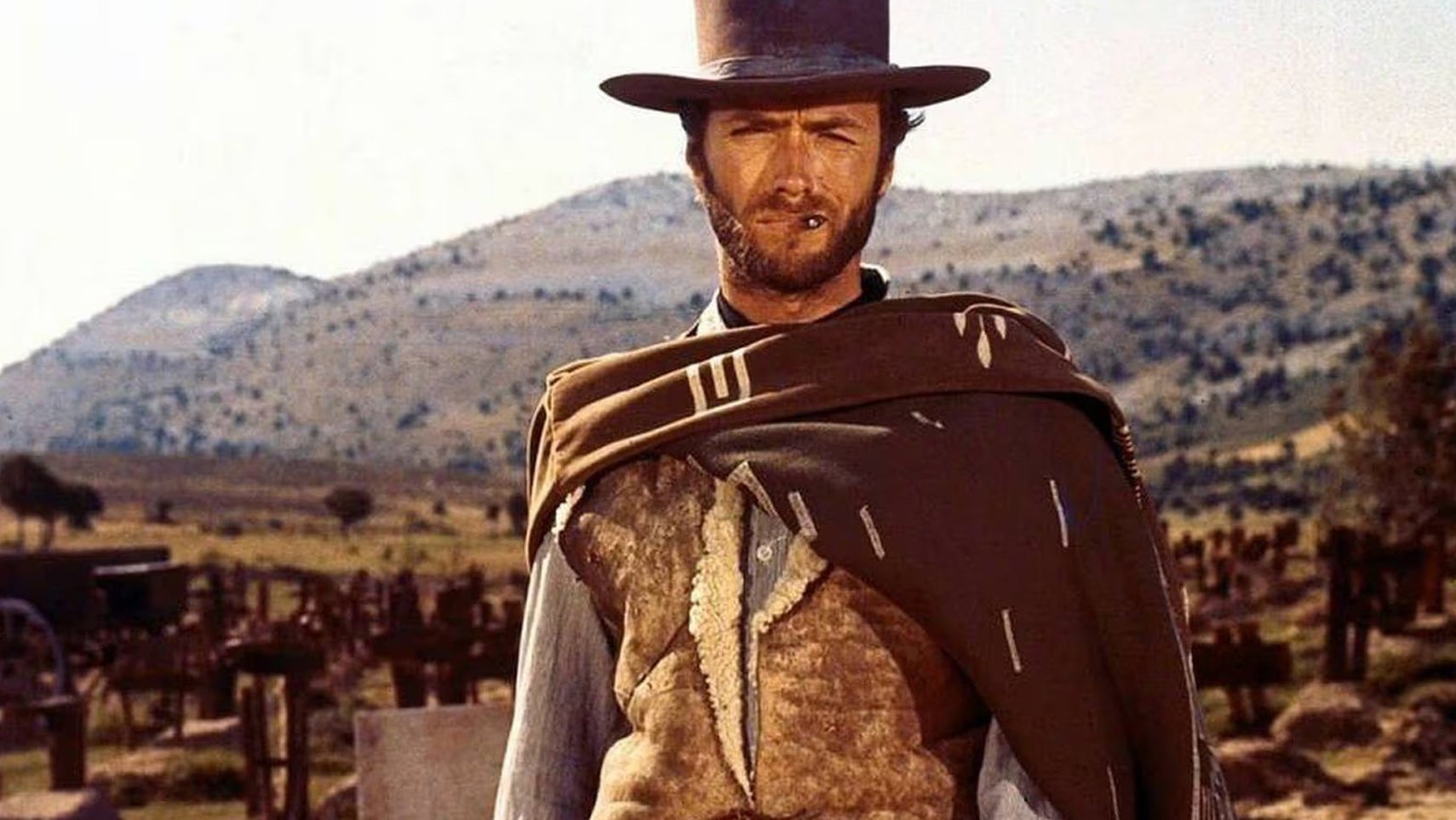 Clint Eastwood as The Stranger in A Fistful of Dollars, he's stood with a thin cigar in his mouth, cowboy hat on his head, looking sternly at something. - 1