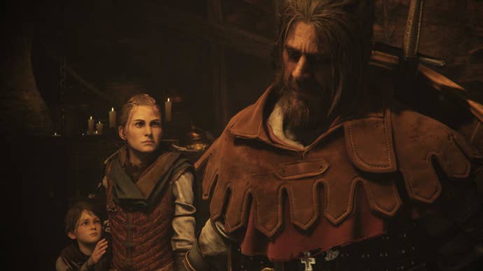 A close-up of Arno, a new soldier companion in A Plague Tale Requiem. He’s got long greying hair tied in a top knot, and a scraggy beard. He’s thick set, with a thick leather hood (that’s down) around his shoulders, and big shield on his back. A young woman (Amicia) and young boy (Hugo) look up at him.