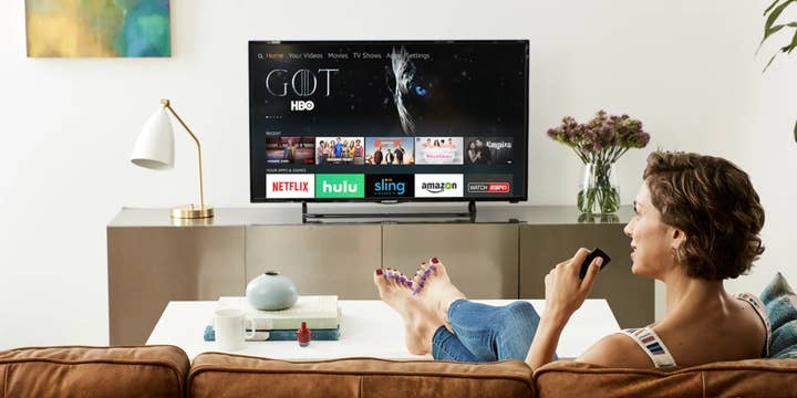 A woman watching TV on the couch talks into an Amazon Fire TV remote control. The Fire TV menu interface is on the TV. She has her feet up on a coffee table with freshly painted toenails. An open bottle of nail polish is on the table next to her feet, but the cap is nowhere to be seen