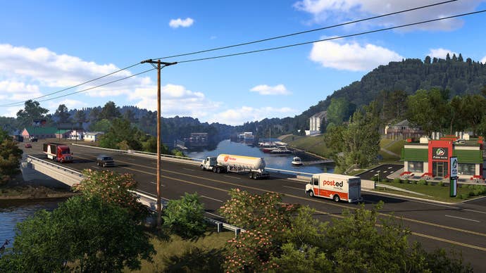 A screenshot of American Truck Simulator’s Arkansas expansion showing vehicles crossing a low multi-lane bridge. A river winding between green, tree-lined hills beneath bright blue skies is visible in the background.