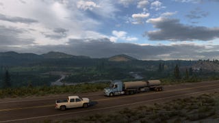 A screenshot of American Truck Simulator's Wyoming DLC showing a truck driving on a road in front of a sprawling vista of river, mountains and trees.