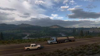 A screenshot of American Truck Simulator's Wyoming DLC showing a truck driving on a road in front of a sprawling vista of river, mountains and trees.