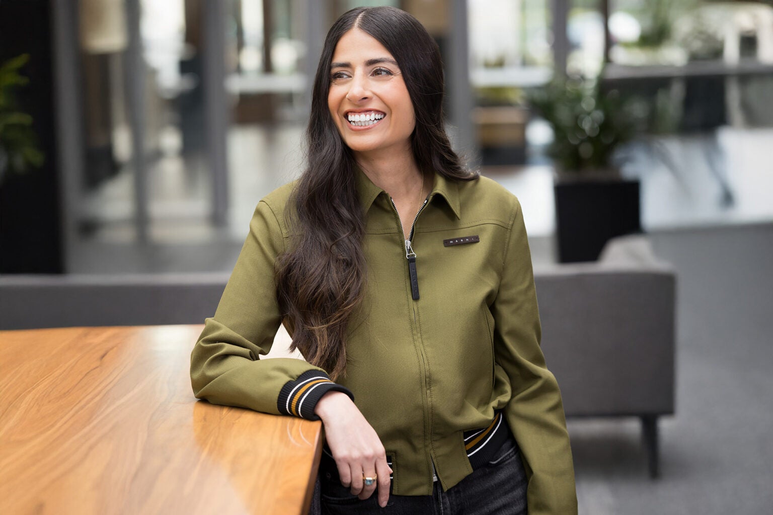 Corporate photo of Asha Sharma, Xbox CEO, wearing a moss green shirt and smiling widely while looking to the side of the camera, in a grey office.