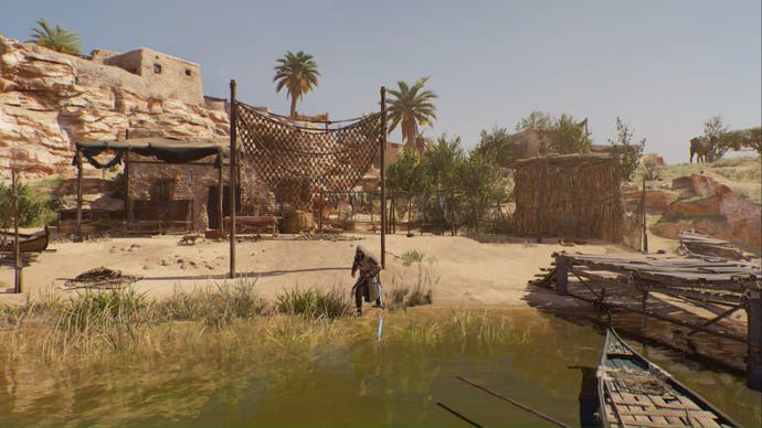 assassins creed mirage, a wide shot of Basim standing on the edge of a river with a boat on the right, fishing nets and fish hanging from wooden poles behind him, plus a house and hut are either side of Basim in the distance.