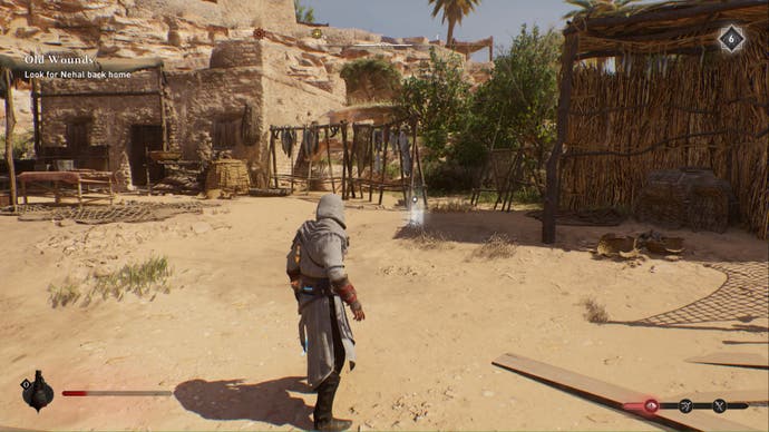 assassins creed mirage, Basim is facing the glowing marker for the left behind enigma treasure location in an abandoned village. There are fish hanging from wooden structures in the distance, a building on the left and a lean-to hut on the right.
