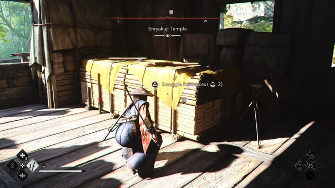 Naoe en Assassin's Creed Shadows se agacha junto a una reserva de recursos dentro de un edificio de madera.