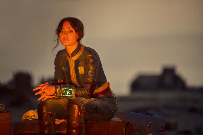 Ella Purnell as Lucy, a Vault-dweller, sits outside lit by the glow of off-screen flames in this screen from Fallout