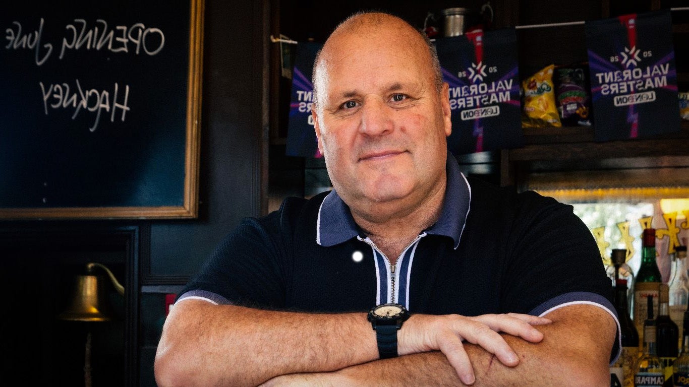 A bald-headed white man folds his arms on the bar of a pub and looks, with a welcoming expression, towards the camera. He'll serve you some beer. I think.