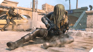 A man with camouflage netting on his head slides across a dusty piece of ground.