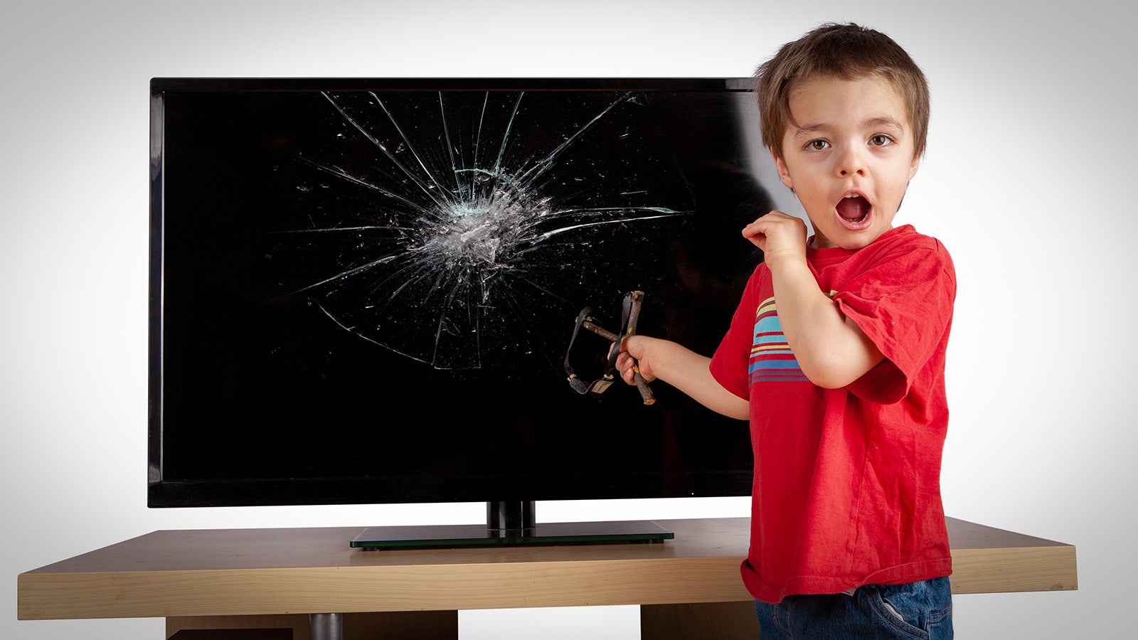 A young child with a grubby face and holding a slingshot looks surprised, as if unexpectedly discovered next to a large, broken TV. Oh naughty naughty Timmy, what did you do?