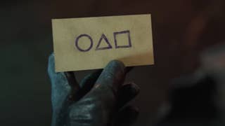 A still from Call of Duty's Squid Game teaser trailer showing a gloved hand holding a small envelop with circle, triangle, and square symbols on the front.