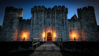 A squat, robust-looking castle close up and at night. Sconces light the wooden door and walls to either side of it. A small wooden footbridge leads over to the entrance.