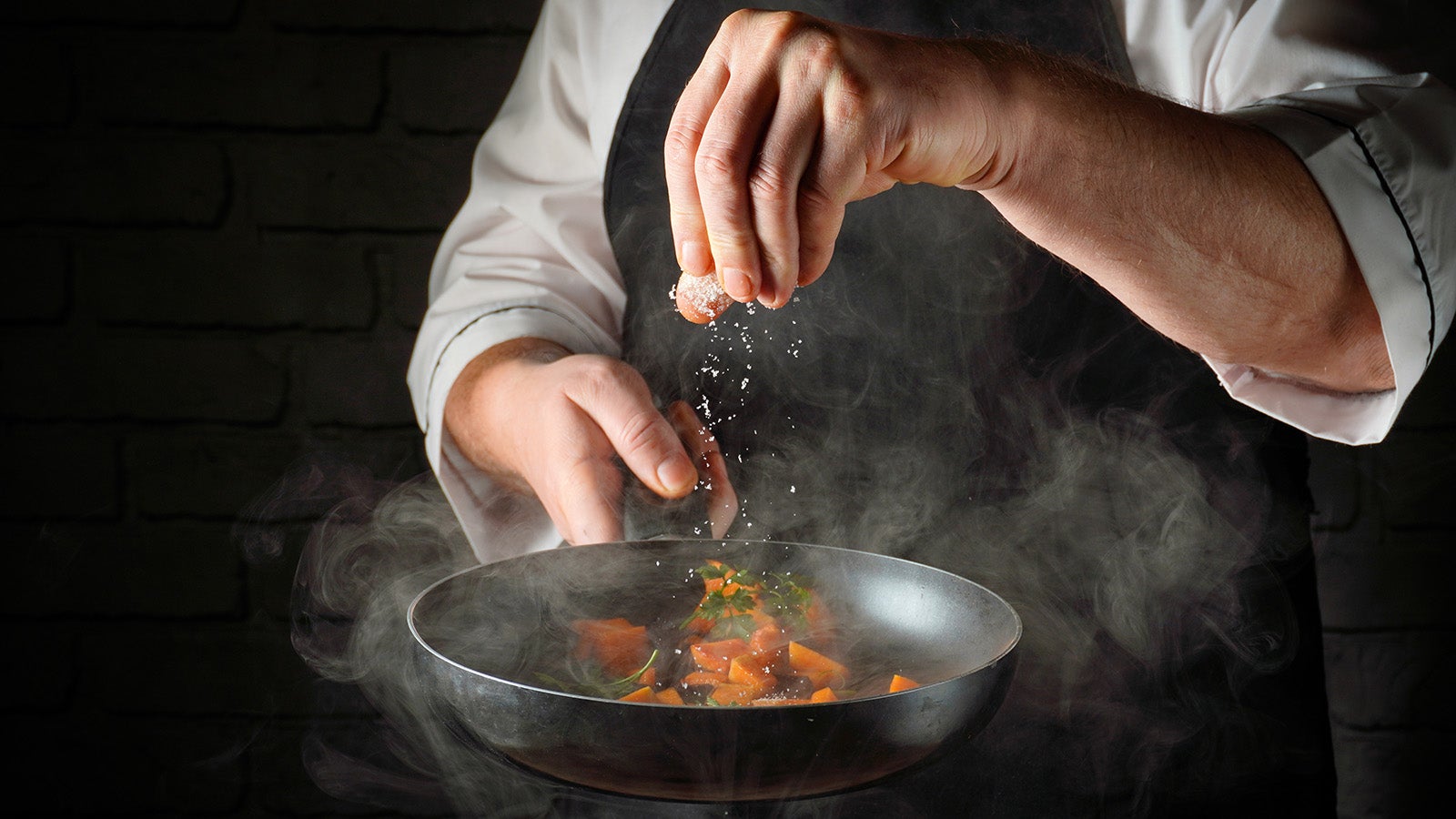 A close-up of a chef in chef whites flipping a small pan with one hand while crumbling some salt into it with the other. There are some orange chunks in the pan. Maybe it's salmon? I hope the chef knows.