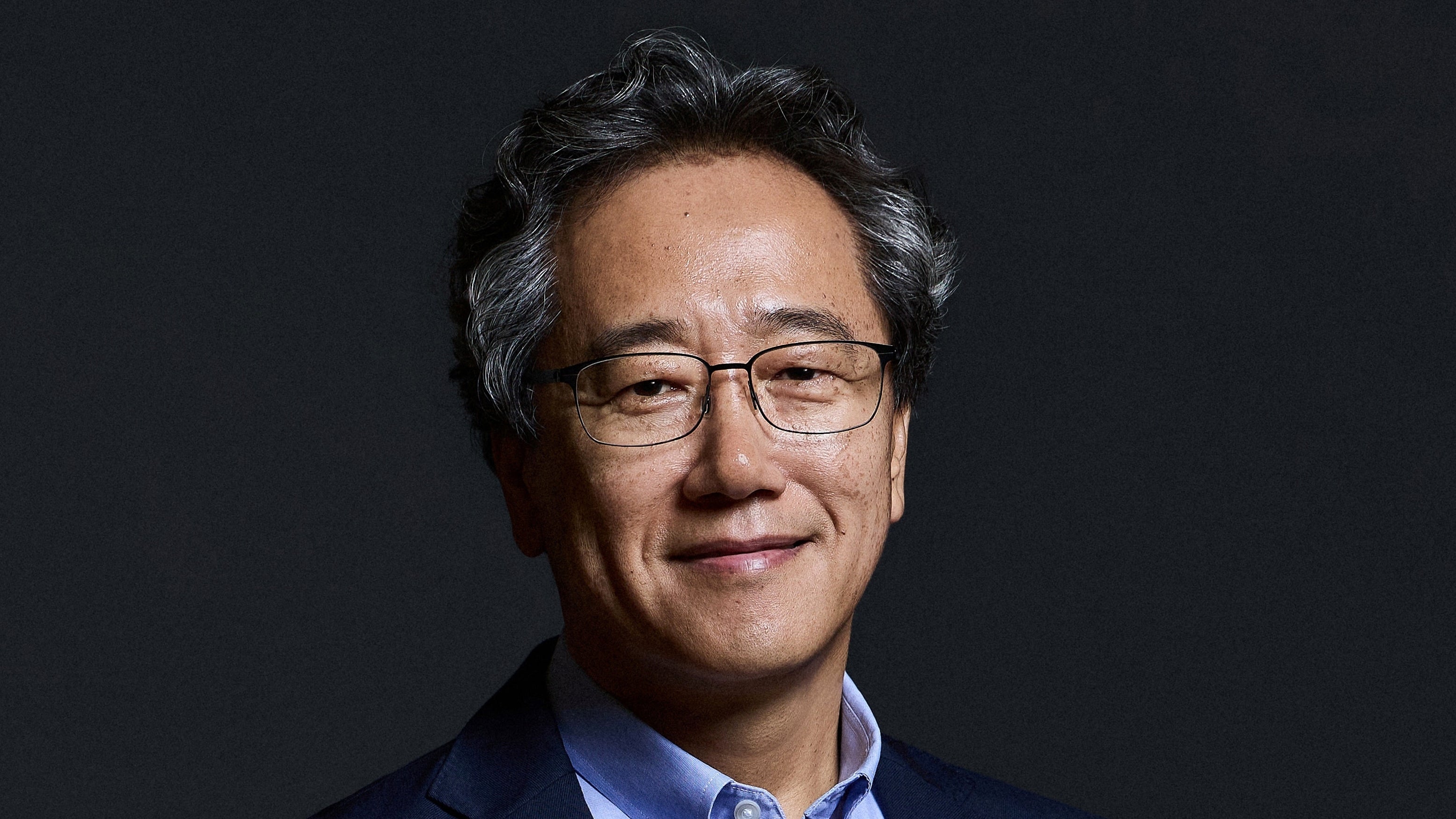 Close-up portrait of a man with salt-and-pepper curly hair, wearing glasses and a blue collared shirt under a dark suit jacket.
