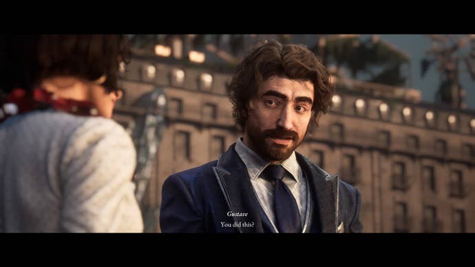 A smartly dressed man in blazer and tie, with a thick moustache and stubbly beard, looks towards a woman in a white polka dot shirt with a kind expression on his face.