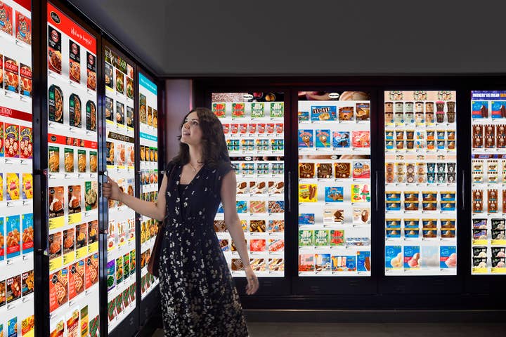 A woman in a store freezer section where all the cooler doors are actually TV monitors showing pictures of the products inside. The woman looks up at some frozen dinners and reaches for the cooler's handle