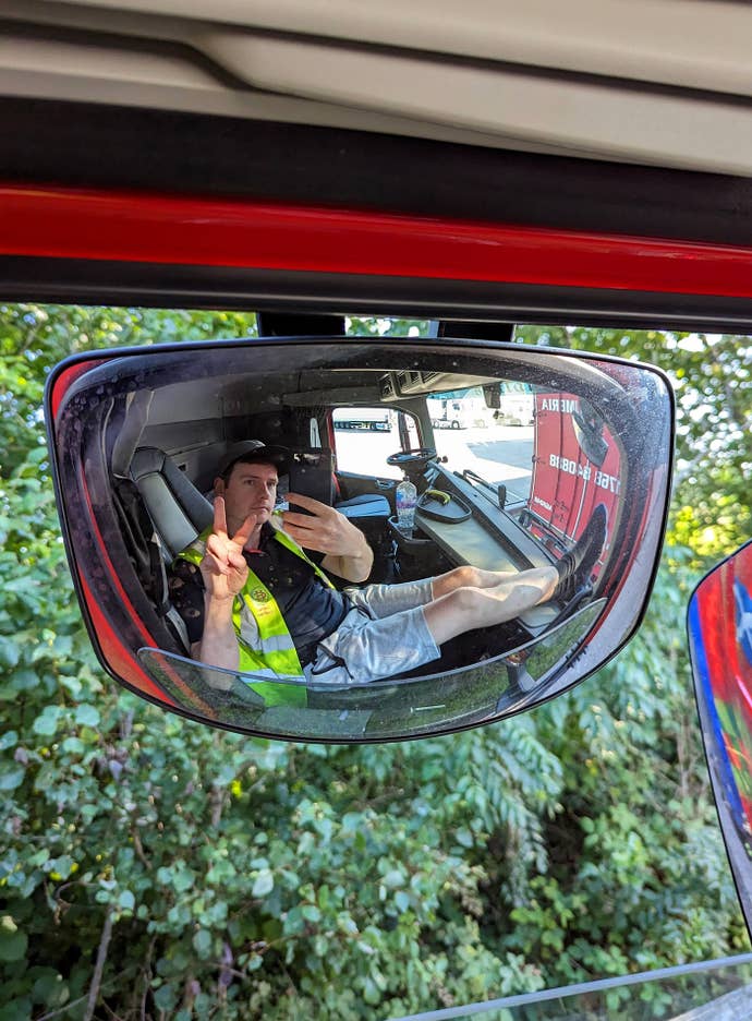 A side-view mirror reflected photograph of a person sitting in a truck cab posing for the camera. That person is Craig Wilmott, and they’re in the truck they drive.