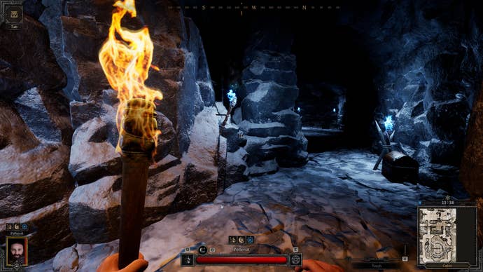 A first-person screenshot inside a rocky cavern dungeon. The player character’s hand can be seen holding a flaming torch. It’s dark and cold in there.
