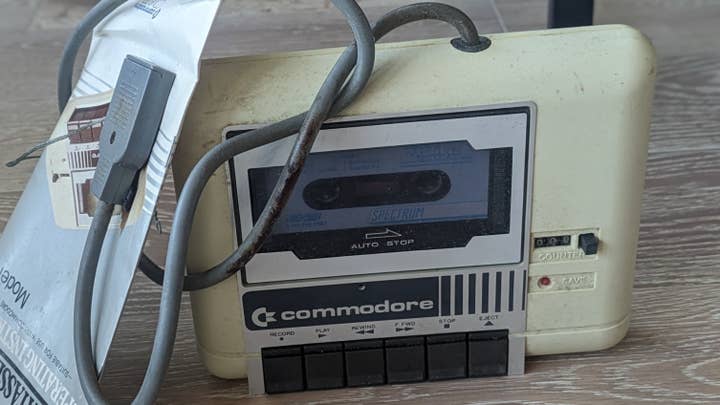 A picture of an old Commodore Datasette tape player. It is scratched and a bit dirty. The rubber on the power cable has started to rot. A crumpled instruction manual leans up against it