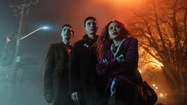 The main cast of Dead Boy Detectives, two young men and a woman, stood in a foggy area, streetlamp to the left of them, trees to the right.