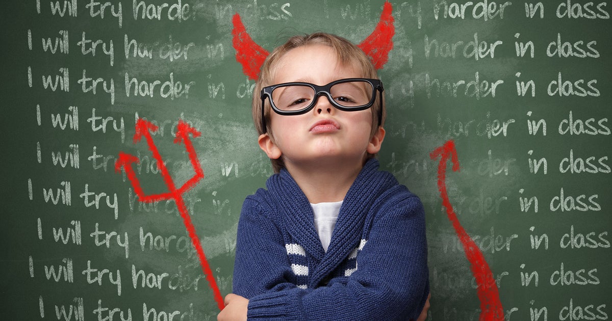 A young boy with fair skin stands with arms crossed, chin up, doing a 'confront me' pose. Someone has drawn devil horns and a trident and a tail on him. Behind, there is a blackboard saying "I will try harder in class" multiple times on it.