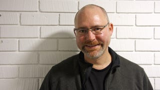 A photo of Geralt voice actor Doug Cockle, standing in front of a white brick wall.