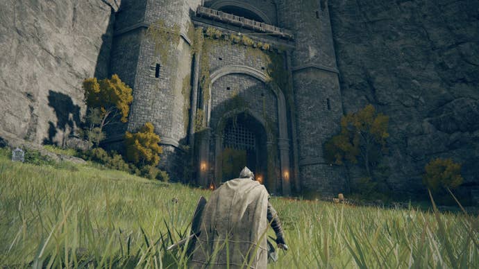 An armoured character wearing a cloak in grass crouching outside of a castle’s entrance in the Limgrave area in Elden Ring.