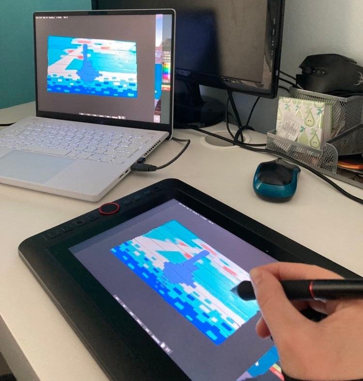 A laptop on a table and a graphics tablet connected to it. A hand holding a pen belongs to the person we can&rsquo;t see who&rsquo;s drawing a picture in the game Rust. It&rsquo;s a blue and colourful scene that appears to be being painted - possibly of a lighthouse. I bet it&rsquo;ll sell for fortunes.