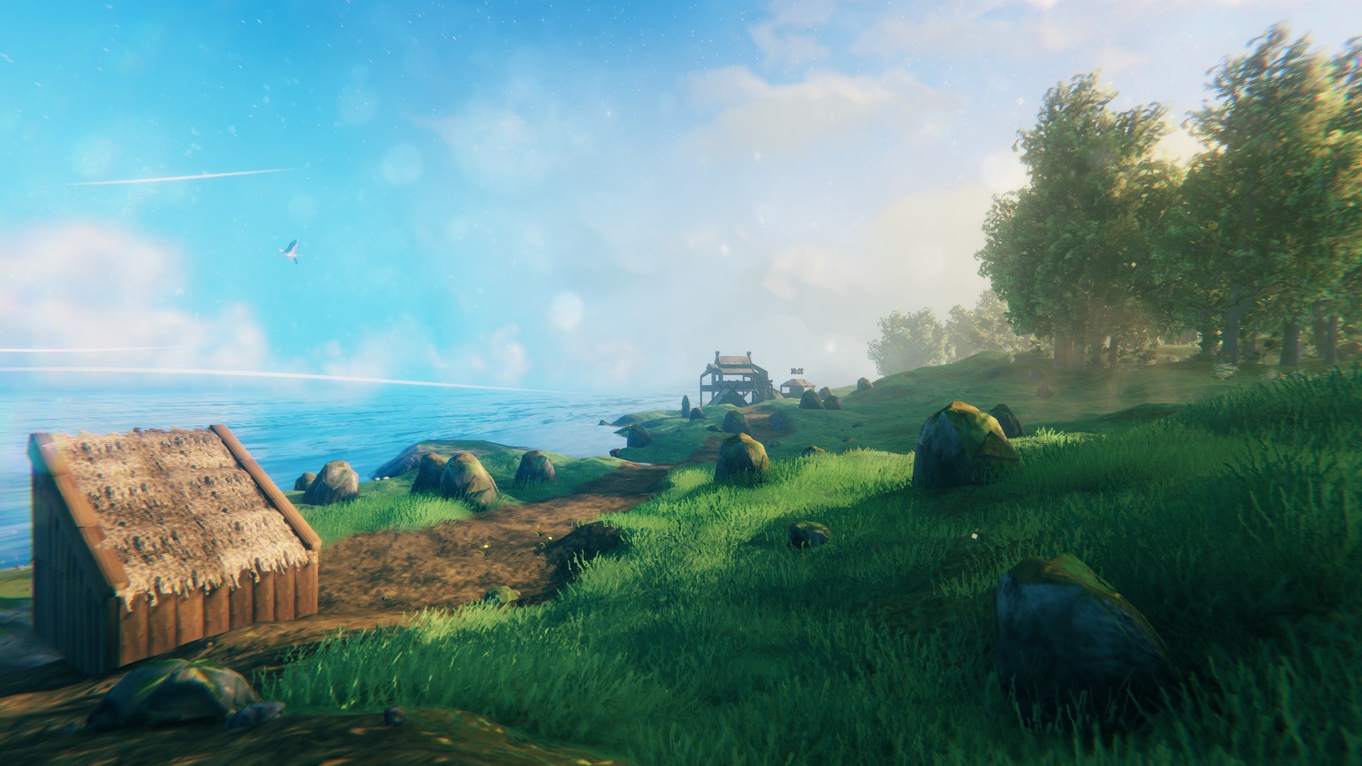 A coastal Valheim scene. Bright blue skies and water, and green grassy fields and tries. In the distance, there&rsquo;s a log cabin house that Emma has built. Leading to it is a quaint little road.