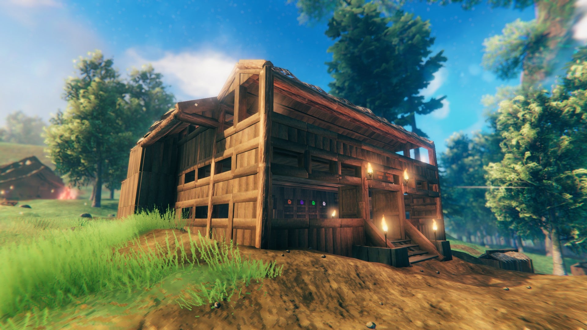 A large, multi-story log cabin in a lightly forested and grassy area. Some torches burn at the top of a small flight of stairs. It&rsquo;s the headquarters of Emma&rsquo;s Valheim real estate Hutenheim Group.