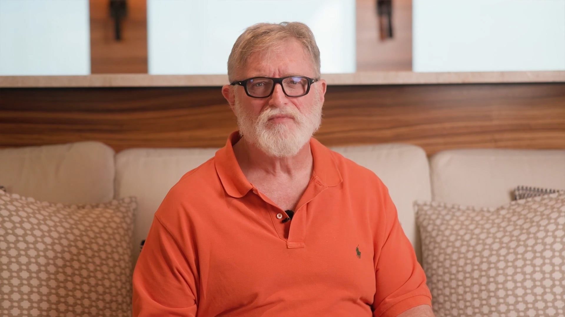 Gabe Newell, a man with grey hair, a white beard, and glasses wearing an orange shirt, is sat on a sofa looking into the camera. - 1