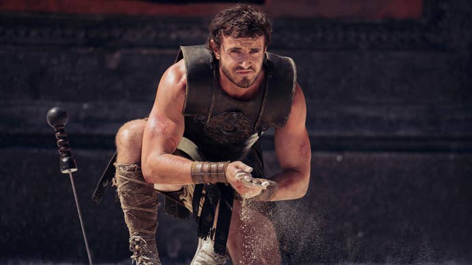 Paul Mescal as Lucius in Gladiator 2, he’s crouched down wearing armour, sword stabbed in the ground, rubbing sand into his hands.
