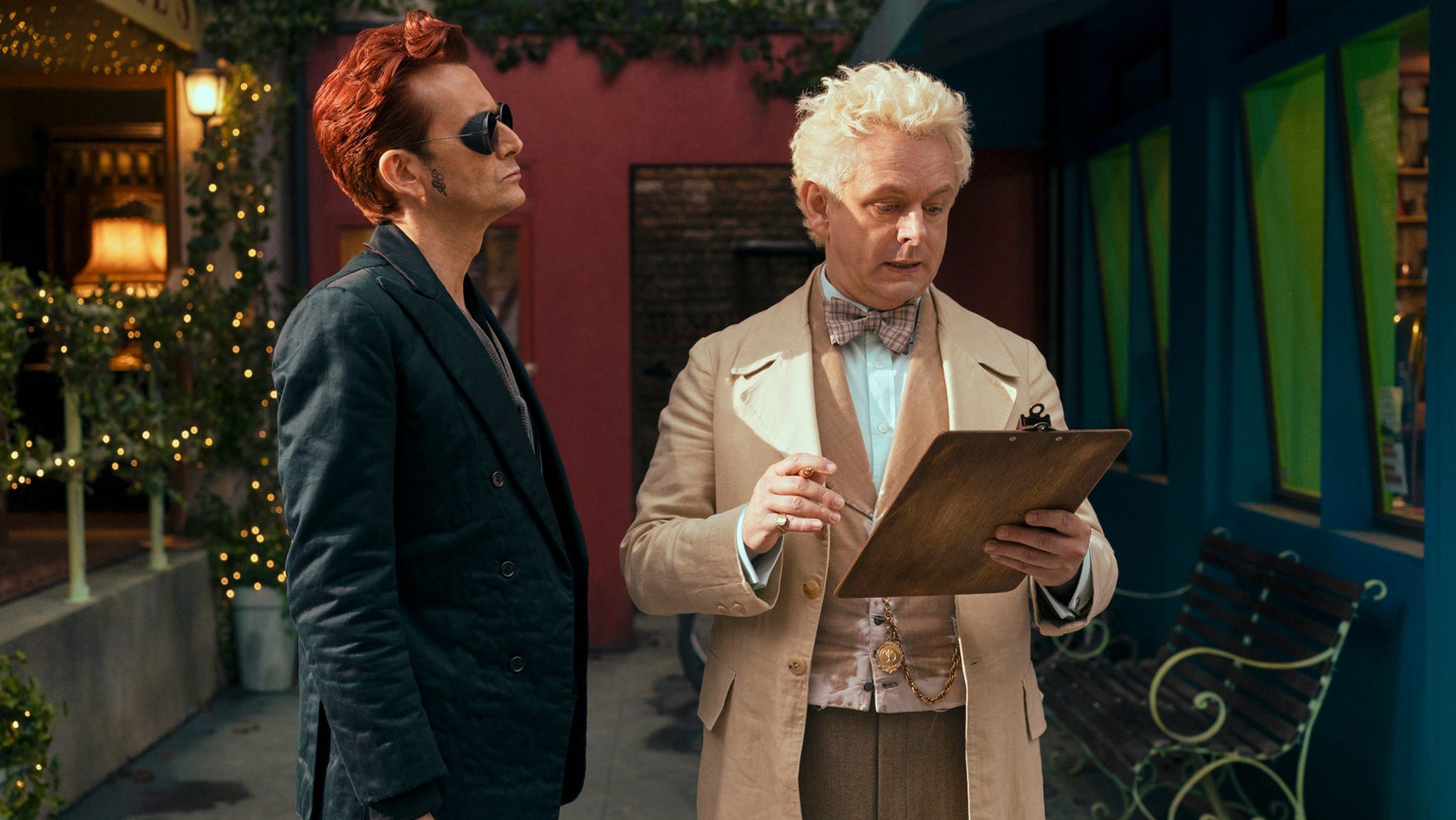 Still from Good Omens showing Crowley, a man with short red hair and dark sunglasses, and Aziraphale, a man with bright blonde curly hair holding a clipboard. - 1