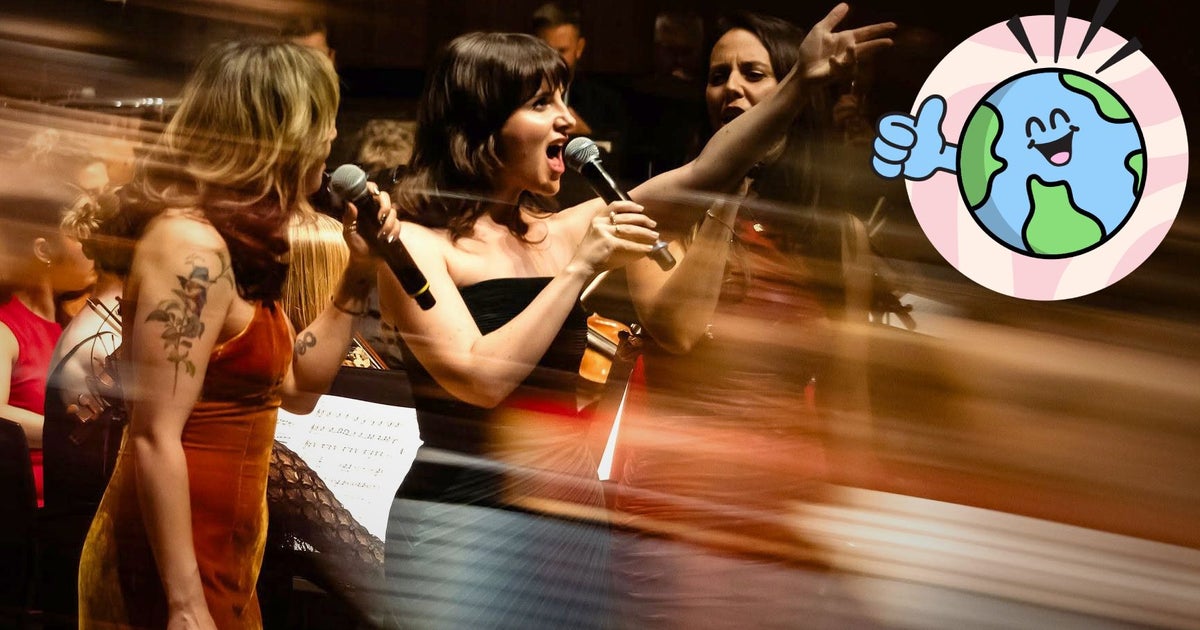 Three singers stand on stage in front of an orchestra, as the central singer raises their hand and belts out a presumably show-stopping note. The edges of the photo are blurred to give it a slightly surreal feel, and a happy, thumbs-upping globe has been overlaid in the bottom co