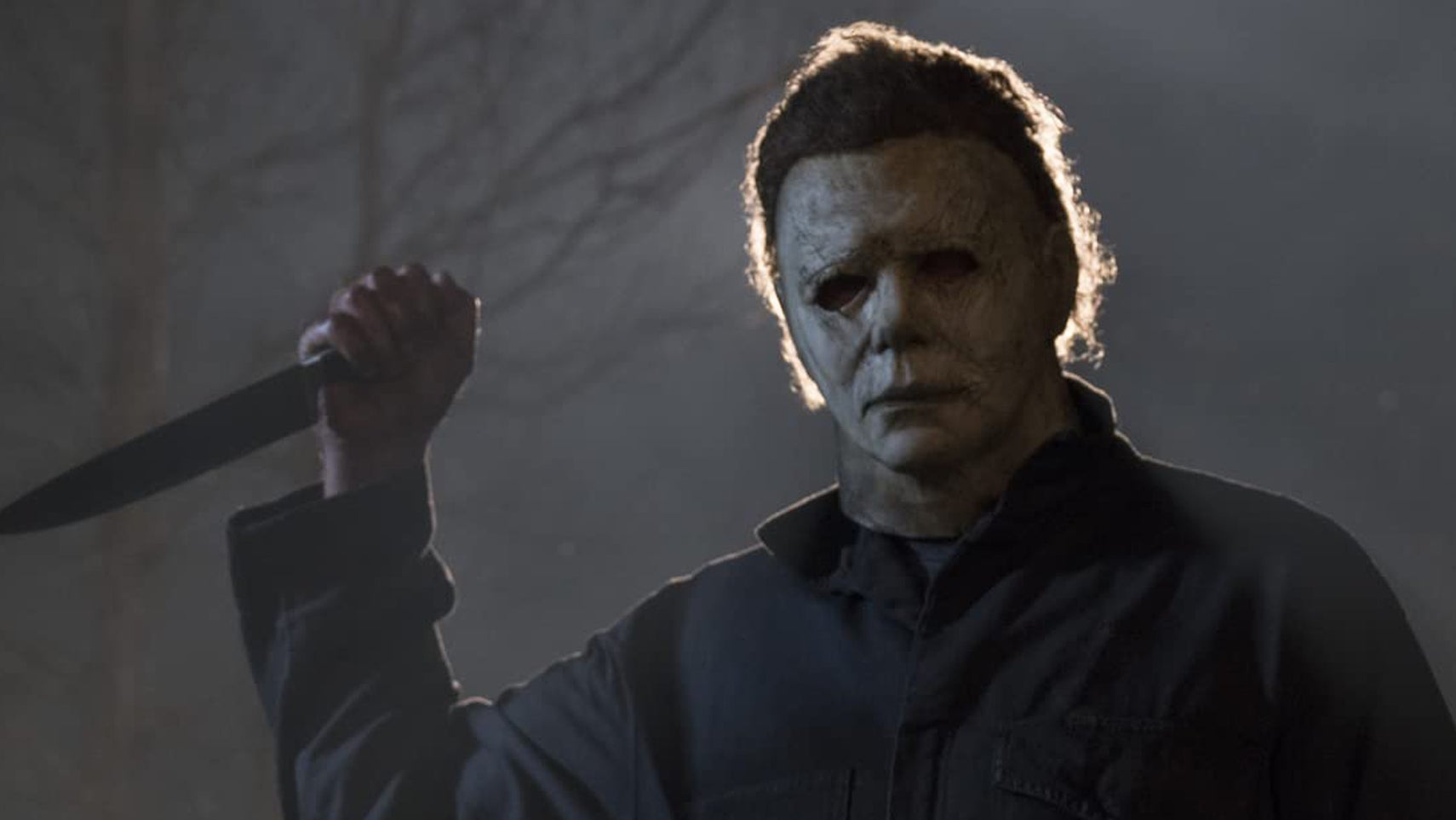 Michael Myers in the original Halloween, he's holding up his classic kitchen knife, it's dark, a light shining behind his head. - 1