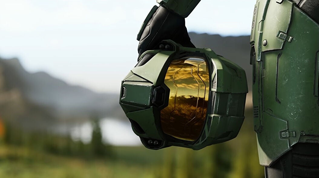 A promotional Halo Infinite image showing a close-up of Master Chief's helmet held by his side while a lush landscape of lakes and mountains is visible in the background.