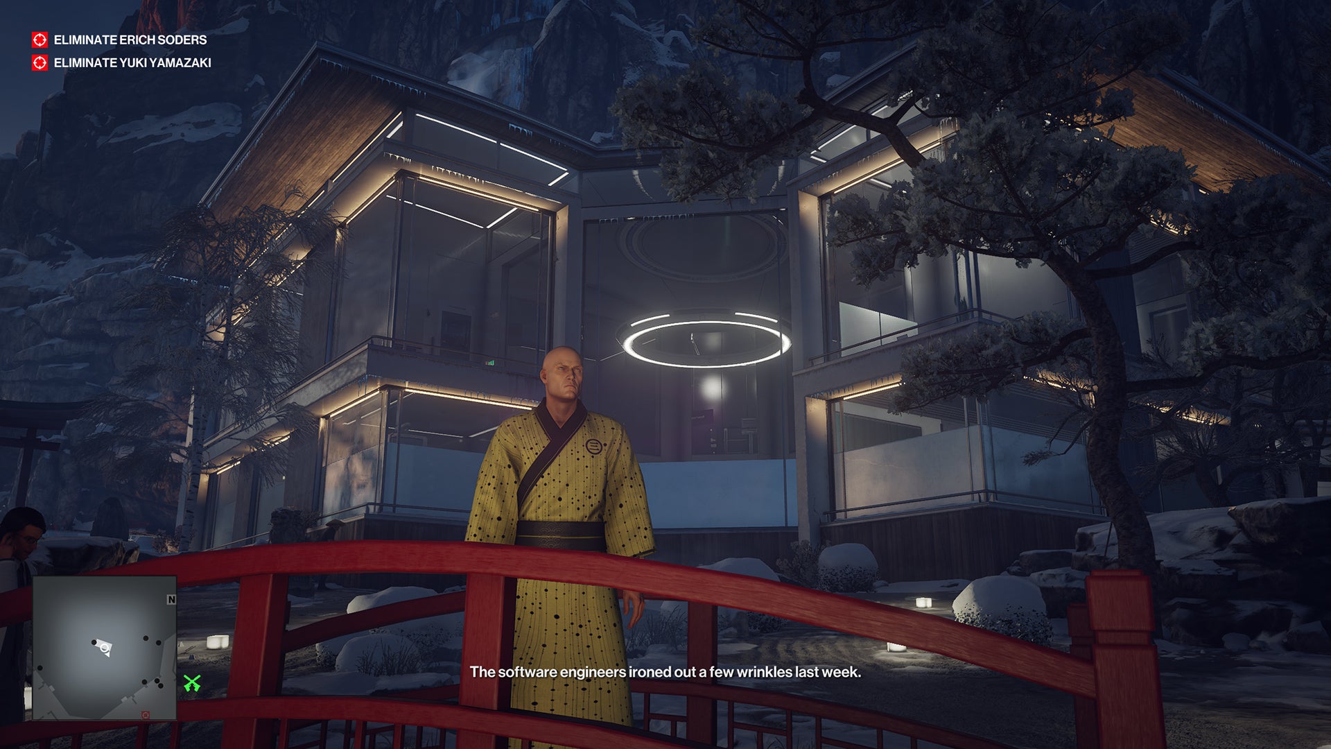 Ian Hitman stands on a bridge in the Hokkaido level. Behind him, the glass walls and soft lights of the posh mountain spa contrast with the dark rock of the mountain around it.