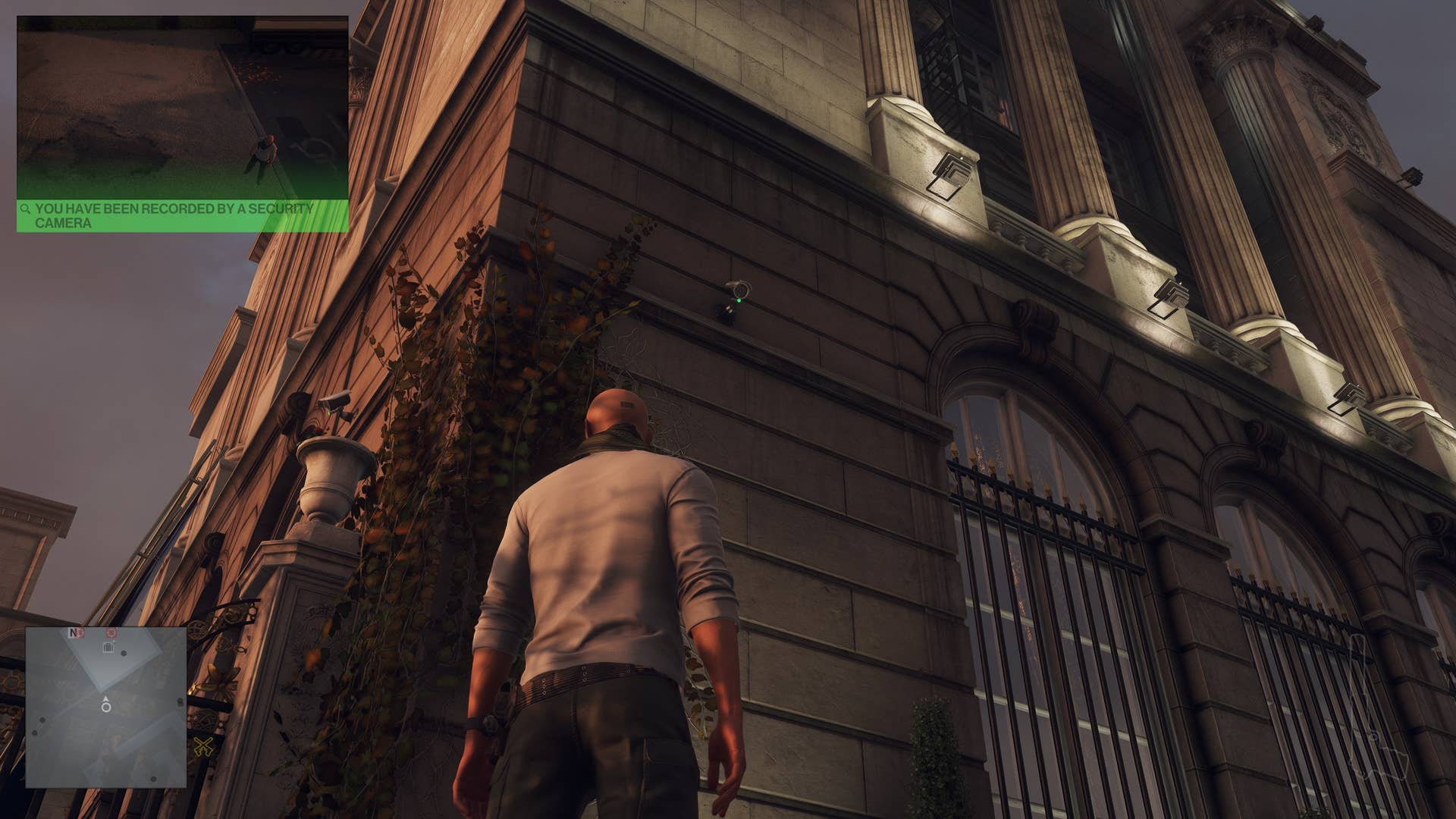 Agent 47 looks up at two security cameras on the side of a large, opulent building.