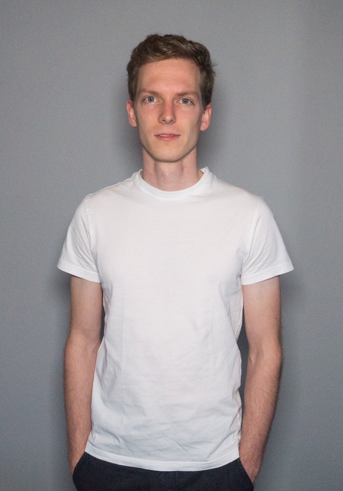 A portrait of Philip Stollenmayer, with short hair and an enigmatic smile. He is wearing a white T-shirt and has his hands in his pockets.