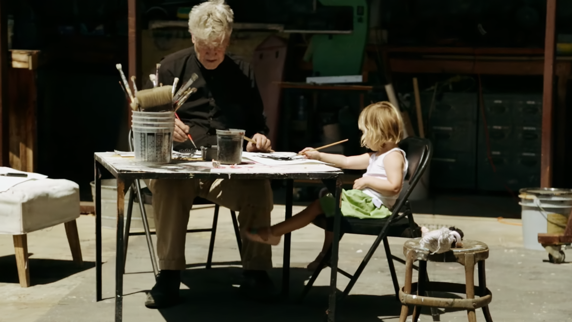 David Lynch and a child sit at a table filled with art supplies working on some art in this screen from the documentary, David Lynch: The Art Life.