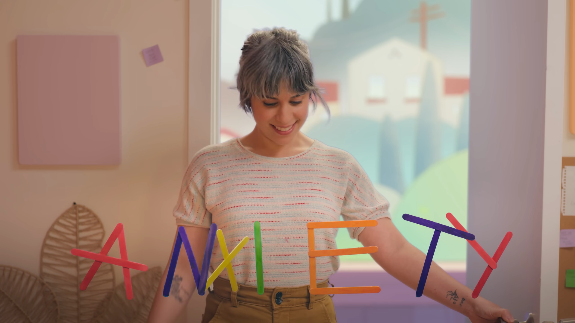 Ashly Burch stands in a brightly lit room with the word ANXIETY spelled out with colourful lollipop sticks floating in the air in front of her