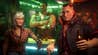 Three gruff-looking people at a neon-lit bar turn to face the camera. One is an older lady, one is a burly barman with their arms crossed, and one is a chunky leather jacket-wearing, broad-faced thug of a guy, with a beer in their hand. It's Jackie Welles from Cyberpunk 2077.