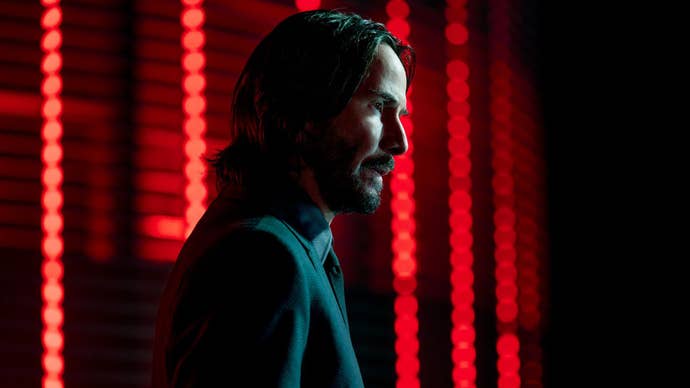 John Wick is stood, it’s dark, his face partially lit by a white light, red lights blurred behind him.
