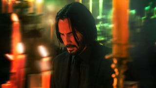 John Wick is stood in a room with candles burning around him, he's looking down solemnly.
