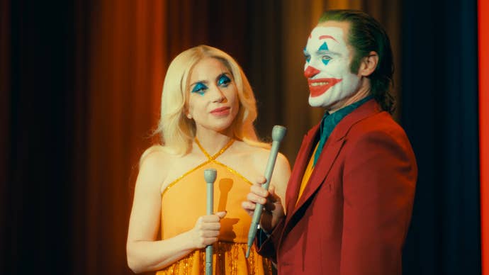 Joaquin Phoenix and Lady Gaga as Joker and Harley Quinn in Joker: Folie a Deux, they are booth stood on a stage, microphones in hand, smiling.