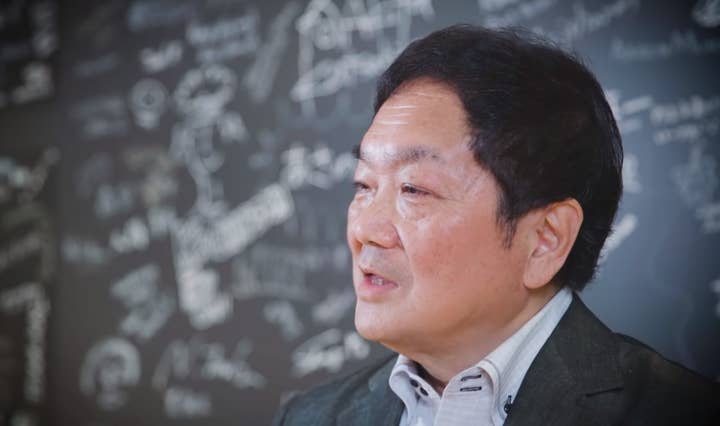Side view of Ken Kutaragi from the shoulders up in a screenshot of a PlayStation 25th anniversary video. He is standing in front of a blackboard with illegible writing on it.