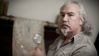 A photograph of a white man with a pointed, Van Dyke white beard, and long white hair neatly swept back over his head, holding a magnifying glass up to some ancient document or other in the background.